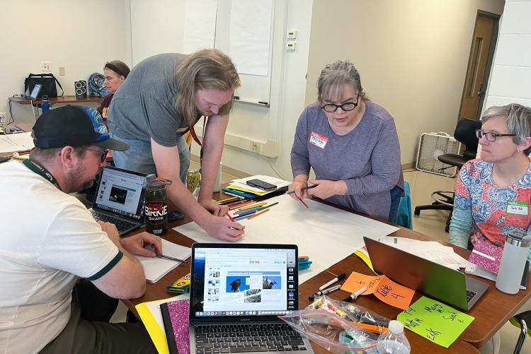 Four people gather around table and draw on a large sheet of paper with colored pencils