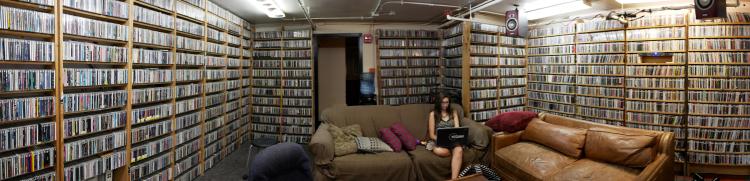 A young woman sits on a sofa, working on a laptop at Radio 1190. She is surrounded by shelves of C.D.s