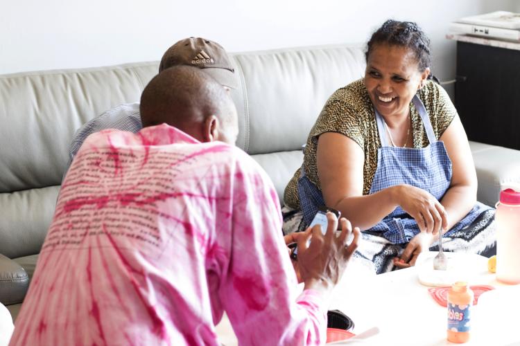 A women with braided hair, an apron, and fork in hand sits on a couch and laughs with two co-workers, whose backs are to the camera, during their lunch break.
