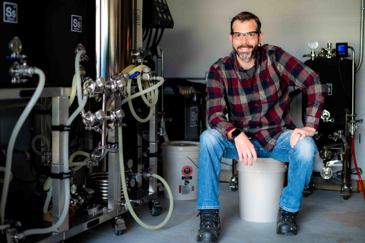 Man in flannel shirt wearing safety glasses sits next to equipment with lots of tubes