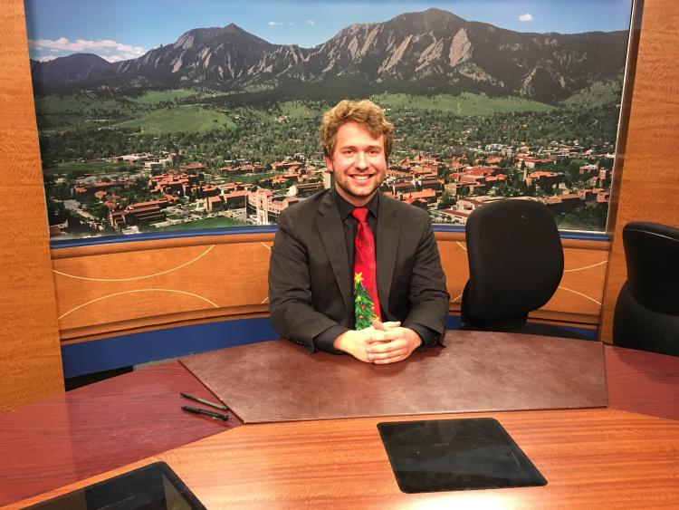 Steve Nelson sits dressed in a black suit with a red tie with a Christmas Tree in front of a backdrop mural of the Fla