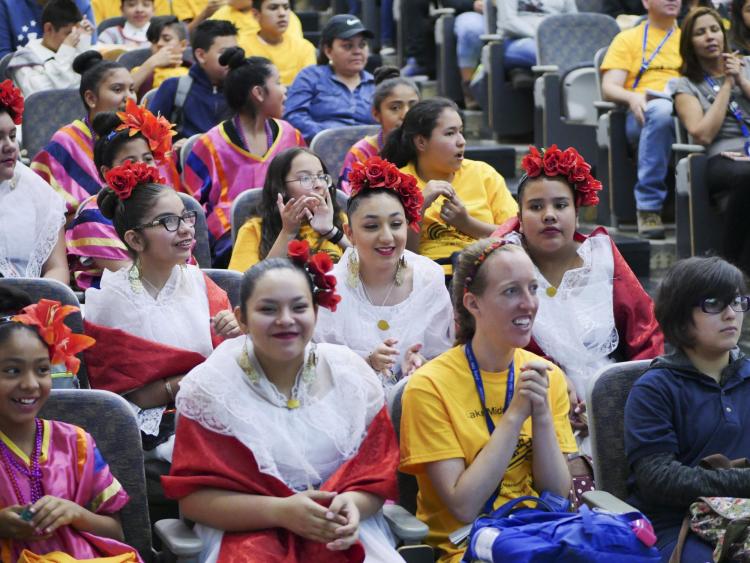 Students donning traditional Spanish dress, sit in auditorium