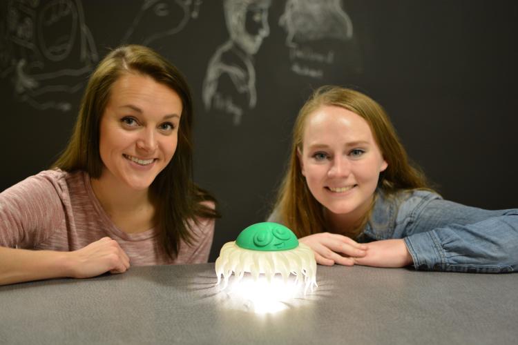 Ariel Wiggan and Sarah Wachter squat behind a gray table with their chest and head above it. Between them is their invention, Tentacles of Light, a lamp which resembles a squid, which has a green head with a face on top and white legs that hang down.