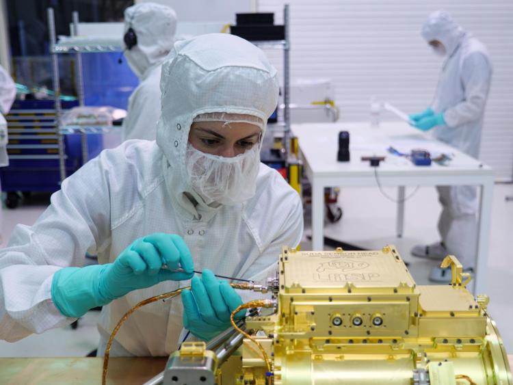 Woman in clean room bunny suit and wearing a mask uses a tool to make an adjustment on a gold instrument
