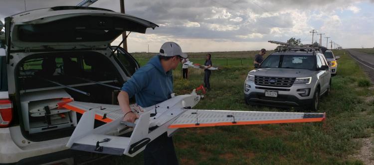Man lifts drone with fixed wings out of the trunk of an SUV while other scientists work in background, and storm clouds brew above