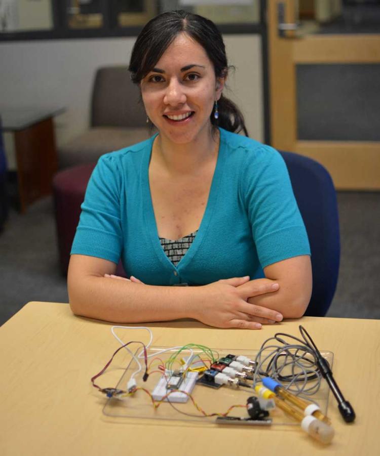 Fuji Robledo, dressed in a light blue shirt with her arms crossed, sits in front of a desk that shows the portable water monitoring system she developed. The device is about the size of a large book and has wires and probes coming out of a clear board.