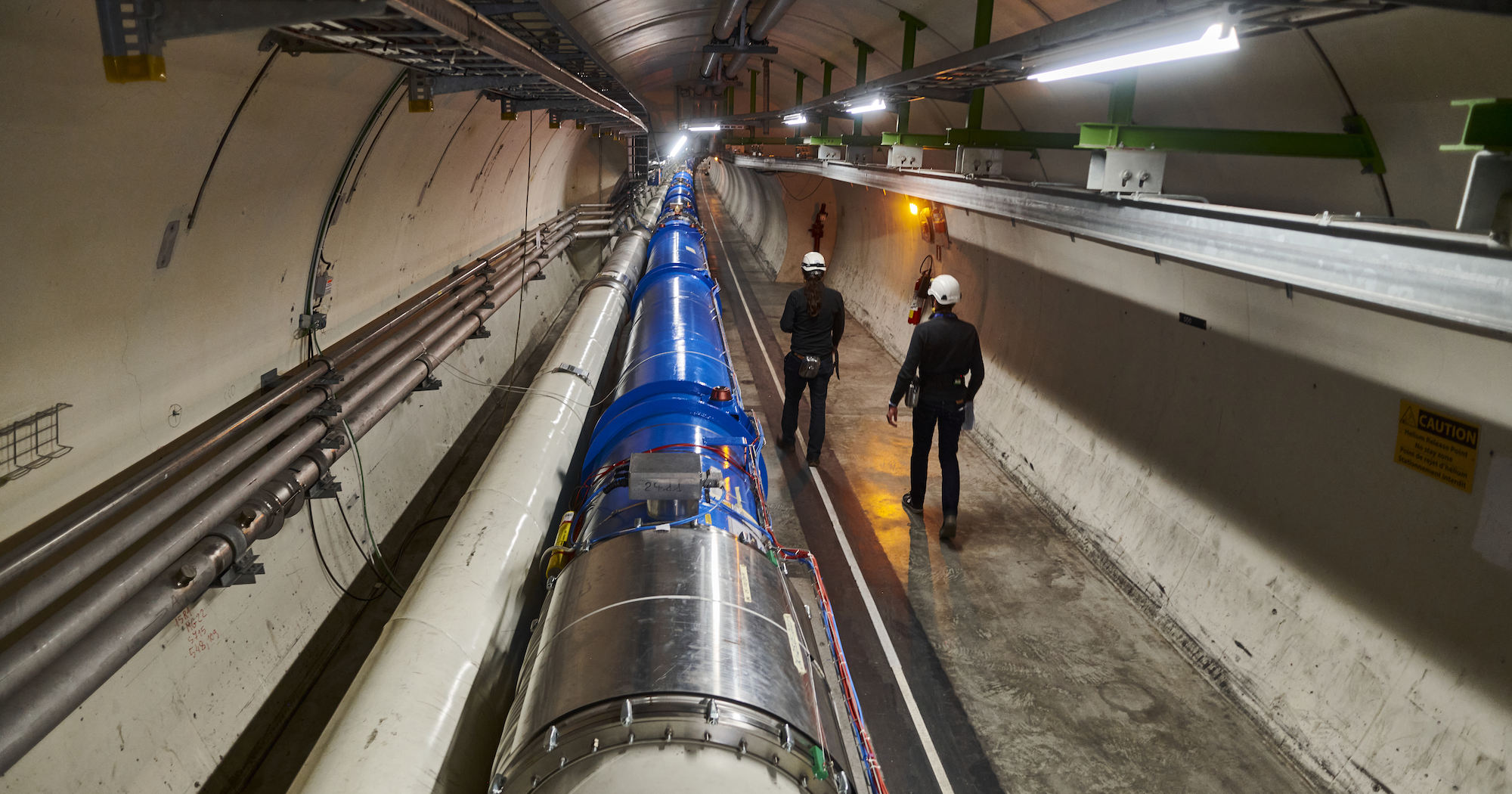 Two people walk beside scientific equipment that stretches down a tunnel