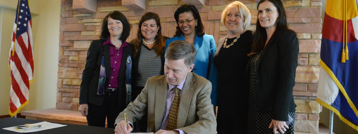 Gov. John Hickenlooper signs bill with people looking on.