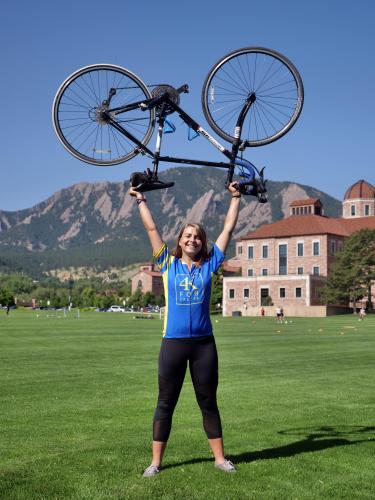 Lauren Gray lifts bike above her head, with Flatirons in the background