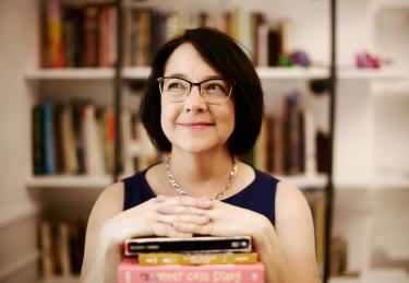 Woman crosses hands over a pile of books with a bookshelf in the backgroundbra