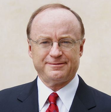 A photo of a white man looking directly at the camera, wearing a blue suit, white shirt and red tie.