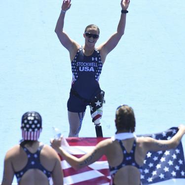 Melissa Stockwell, in stars-and-stripes track wear, with arms extended above her head, faces two people holding an American flag. (Wikimedia Commons/Tânia Rêgo/Agência Brasil)