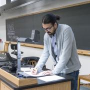 A faculty member, standing near a table and projector, appears to be teaching.