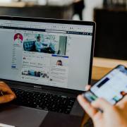 hand holds a smartphone while the other hand scrolls through a facebook profile on a laptop