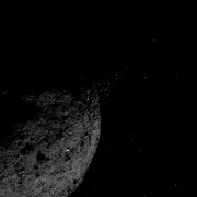 Image of the asteroid Bennu with small bits of material jumping off into space.