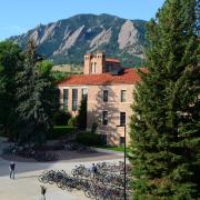 The CU Boulder Hellems building is brightly lit by the sun, with evergreen trees flanking it and the Flatirons and a blue sky in the background.