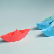 red paper boat leads a pack of blue and green paper boats