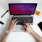 a person working on a laptop with a notebook, pen, phone and an orange nearby