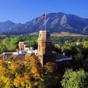 A photo of the Old Main tower with a U.S. flag on top, surrounded by colorful tree leaves and the Flatirons.