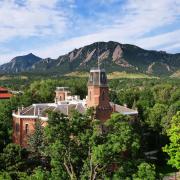 Old Main is seen in an aerial shot, surrounded by the green leaves of summer and the Flatirons in the background. (Casey Cass)