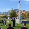 On a grassy lawn, a man speaks at a podium with several others sitting in chairs beside in. In the background, a metal tower reaches up more than 20 feet