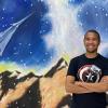 Dezell Turner stands with his arms folded in front of a mural showing mountains, a starry sky and a spaceshift flying above
