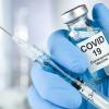 Hand wearing blue glove holds a syringe and a glass vial with a label that reads "COVID 19"