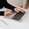 Arms in a black sweater extend to a laptop keyboard, with a phone and pen on the table. (Photo: Pexels)