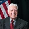 The late President Jimmy Carter in suit and red tie with an American flag in the background