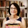 Woman crosses hands over a pile of books with a bookshelf in the backgroundbra
