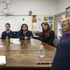 School children in blue blazers sit around a large table speaking to woman in a blue pantsuit