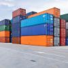 More than a dozen shipping crates stacked on dry ground, likely in a port. The crates are blue, orange, green and gray.