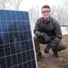 Man kneels on the ground next to a large solar panel with snow in the background