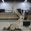 A mechanical X-ray bomb disposal device, which looks like a metal arm with a joint mid-way up, sits on the ground of a lab including a long computer desk in the background.