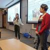 Emily Jerris and Annalise Cabra stand in front of two large screens presenting slides, and address a seated audience in a classroom setting.