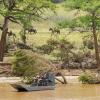 Air boat moves down swollen river with people riding horses in the background