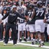 Buff football players walk on the sideline while Coach Deion Sanders extends his left hand to greet on player in passing. (Glenn J. Asakawa/University of Colorado)