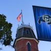 An American flag atop the Old Main tower is seen, with a CU buffalo banner (right) and tree leaves (left) flanking the image.
