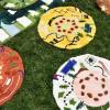 Whimsical plate-like objects in yellow, white and red with geometric and nature shapes rest on green grass.
