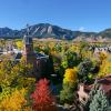 The Old Main building is nestled among trees showing off colorful fall leaves with the Flatirons off in the distance, all under a bright blue sky.