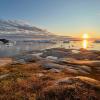 A wide view of shallow water with small clumps of icy snow is seen extending from a rocky shore with a low sun and light stretching clouds in view.