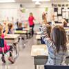 Children, seen from behind, sit at desks and raise their hands in a classroom
