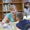 A college student gestures as she sits at a table and works with a young student in what appears to be a library setting.