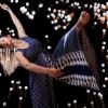 In a horizontal dance position, Erika Randall extends her left leg with a pointed toe, and reaches back with her left arm, round golf-ball size lights glimmering around her with a black background.