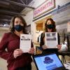 Two women hold up educational materials near a set of cash registers
