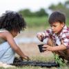 Two children kneel in the grass, scooping piles of dirt into plastic pots