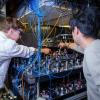 Two men seen from behind lean over a lab table that's covered in machinery, lenses and wires