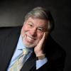 In a portrait with a dark studio background, Steve Wozniak leans forward, propping his face on his hand, smiling into the camera.