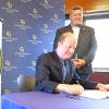 Man sits at table to sign document while another man looks on. A podium bearing the seal of the State of Colorado is in the foreground