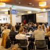 Numerous people sit at large round tables in a conference space, turning their attention to a speaker holding a microphone and presenting from a lecturn. 
